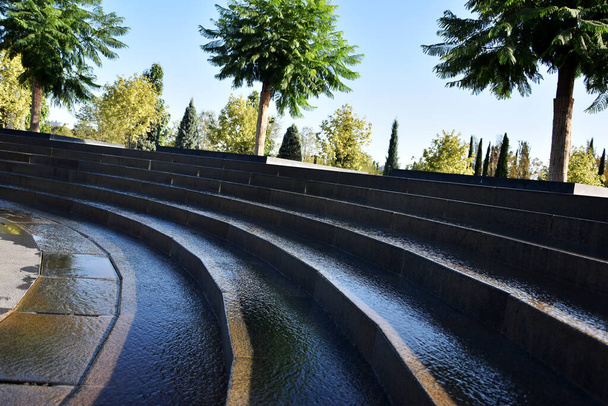 Het water loopt de betonnen trap af, en in de verte groeit een mooie boom. Ontwerp van een modern park met water en bomen. - Foto, afbeelding