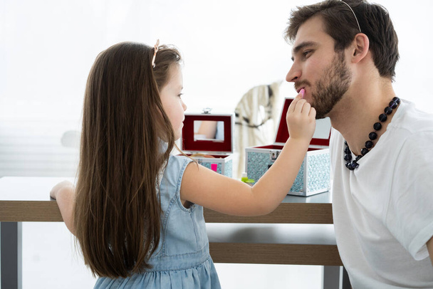Cute little daughter and her handsome young dad in crowns are playing together in childs room. Girl is doing her dad a makeup. - Φωτογραφία, εικόνα