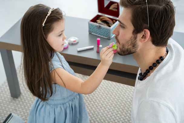 Cute little daughter and her handsome young dad in crowns are playing together in childs room. Girl is doing her dad a makeup. - Foto, immagini