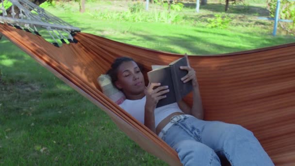 Nuori nainen lukee kirjaa makatessaan riippumatossa metsässä. - Materiaali, video