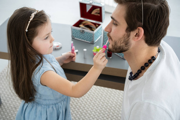 Cute little daughter and her handsome young dad in crowns are playing together in childs room. Girl is doing her dad a makeup. - Foto, afbeelding