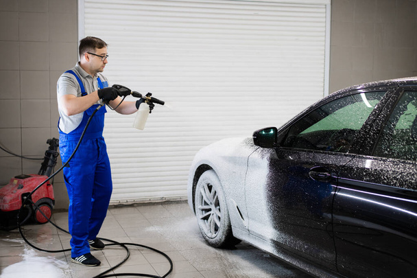Car with turned on backlights standing in car wash service. Worker cover the car with foam with high-pressure sprayer. - Φωτογραφία, εικόνα