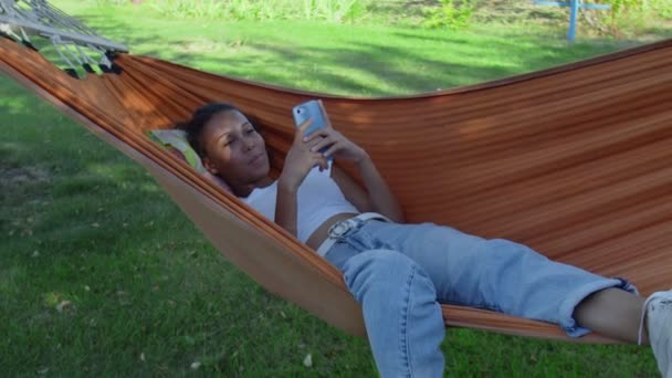 Νεαρή Αφροαμερικανή γυναίκα με το κινητό της τηλέφωνο να χαλαρώνει στην αιώρα. - Πλάνα, βίντεο