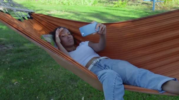 Νεαρή ευτυχισμένη Αφροαμερικανή που βγάζει selfie φωτογραφία με κινητό ξαπλωμένο στην αιώρα - Πλάνα, βίντεο