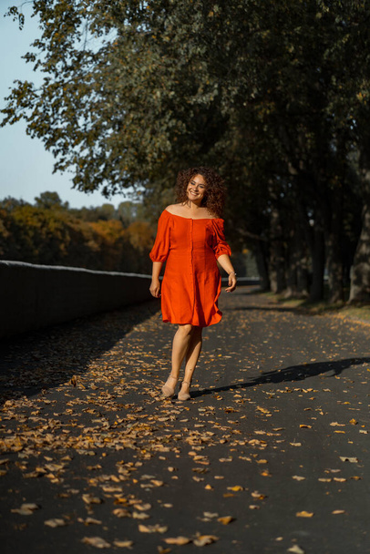 A beautiful woman with curly dark hair in a red dress walks through the autumn city. Autumn mood, yellow foliage underfoot. - Фото, изображение