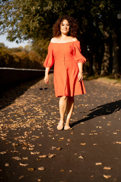 A beautiful woman with curly dark hair in a red dress walks through the autumn city. Autumn mood, yellow foliage underfoot. - Фото, изображение
