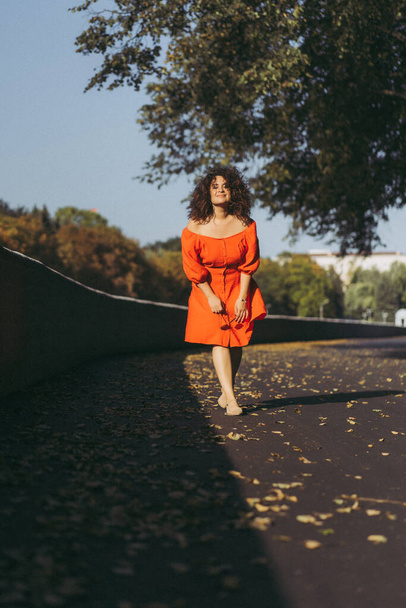 A beautiful woman with curly dark hair in a red dress walks through the autumn city. Autumn mood, yellow foliage underfoot. - Fotoğraf, Görsel