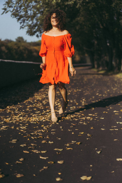 A beautiful woman with curly dark hair in a red dress walks through the autumn city. Autumn mood, yellow foliage underfoot. - Фото, зображення