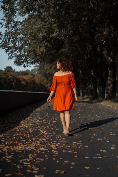 A beautiful woman with curly dark hair in a red dress walks through the autumn city. Autumn mood, yellow foliage underfoot. - Valokuva, kuva