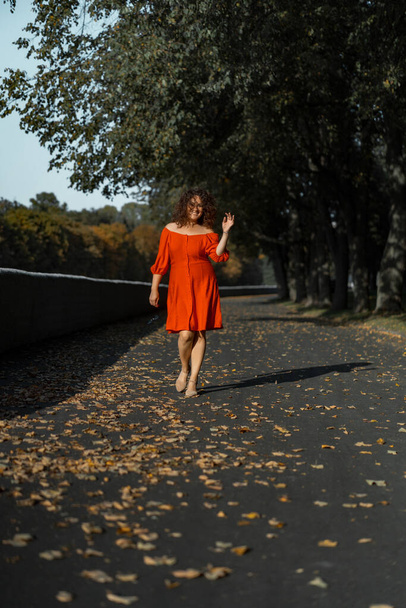 A beautiful woman with curly dark hair in a red dress walks through the autumn city. Autumn mood, yellow foliage underfoot. - Fotó, kép