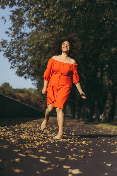 A beautiful woman with curly dark hair in a red dress walks through the autumn city. Autumn mood, yellow foliage underfoot. - Photo, image