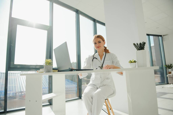 Portrait of smiling young Caucasian woman nurse sit at desk in hospital or private clinic, profile picture of happy positive female doctor GP in white medical uniform at workplace, healthcare concept - Valokuva, kuva