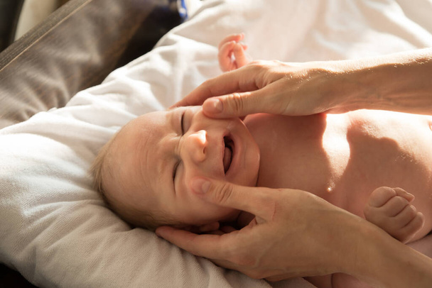 Un bebé llorando tumbado en una cama sobre sábanas blancas mientras su padre le masajea la cara - Foto, Imagen