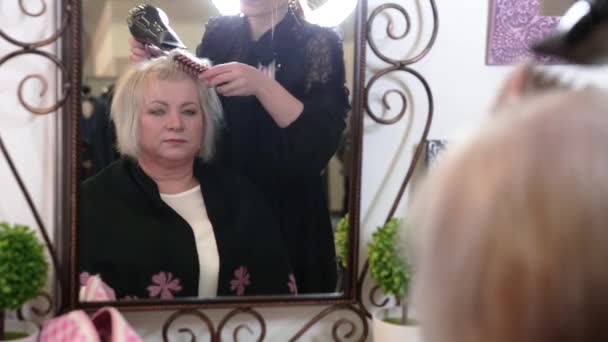 白髪の短い年配の女性が美容室でスタイリングをしています。鏡の前に座っている年配の女性 - 映像、動画