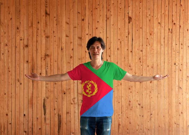 Man wearing Eritrea flag color shirt and standing with arms wide open on the wooden wall background, green red triangle on blue and green triangle with olive branch on gold color. - Photo, Image