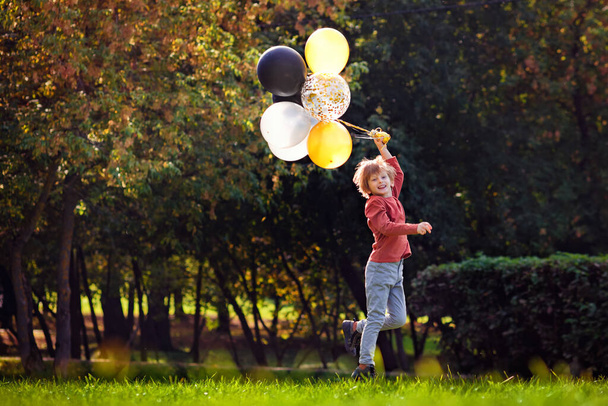 Mladý zrzavý chlapec hraje s partou žlutých bílých černých balónků v parku. - Fotografie, Obrázek
