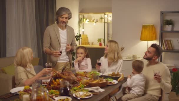 Dojrzały mężczyzna stojący przy stole proponując tosty, a następnie brzęcząc szklanki z rodziną podczas świętowania Święta Dziękczynienia - Materiał filmowy, wideo
