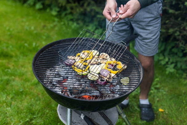 Genç bir erkek, sağlıklı bir parti yemeği için dumanlı bir tat alabilmek için mangal ızgarasının üzerinde sebze pişiriyor. - Fotoğraf, Görsel