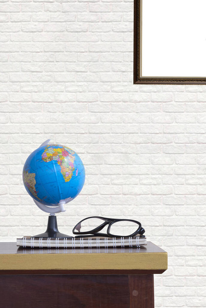 Pequeno modelo globo com óculos e caderno em espiral na mesa de madeira com parte da moldura em branco vintage no fundo da parede de tijolo branco na sala de estar e quadro vertical - Foto, Imagem