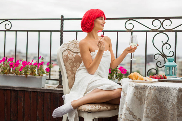 beautiful young female with red hair in a white towel eats strawberries at a table on the terrace - Photo, Image