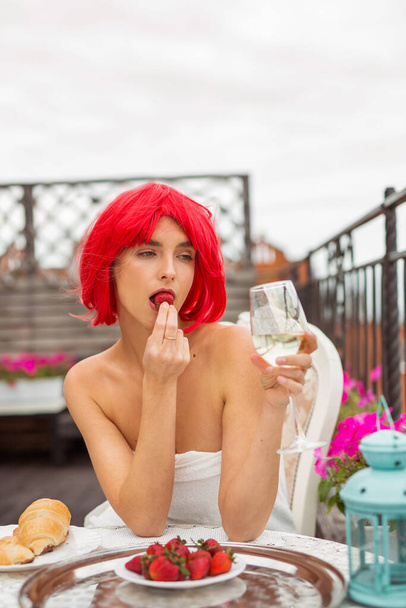 beautiful young female with red hair in a white towel eats strawberries at a table on the terrace - Photo, image