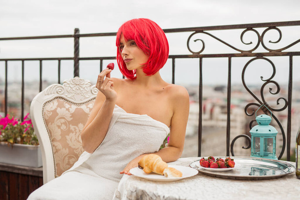 beautiful young female with red hair in a white towel eats strawberries at a table on the terrace - Photo, Image