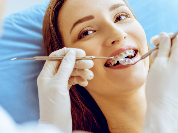 歯科医院で歯科医に検査された歯列矯正ブラケットを持つ笑顔の若い女性。健康な歯と医療の概念 - 写真・画像