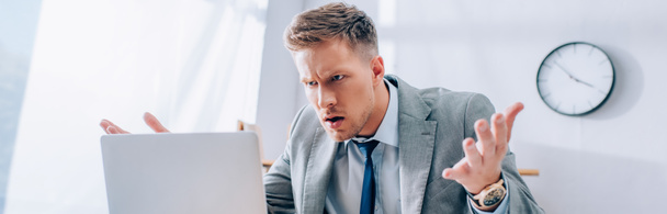 Агрессивный бизнесмен смотрит на ноутбук на размытом переднем плане в офисе, баннер  - Фото, изображение
