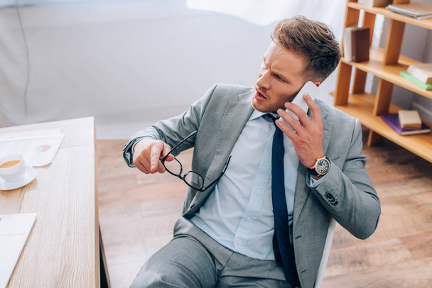 Angry businessman talking on smartphone and holding eyeglasses in office  - Foto, afbeelding