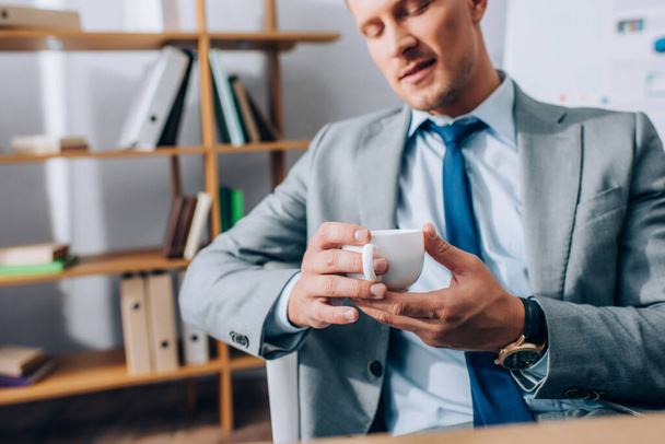 Cropped view of cup of coffee in hands of businessman on blurred background in office  - Фото, зображення