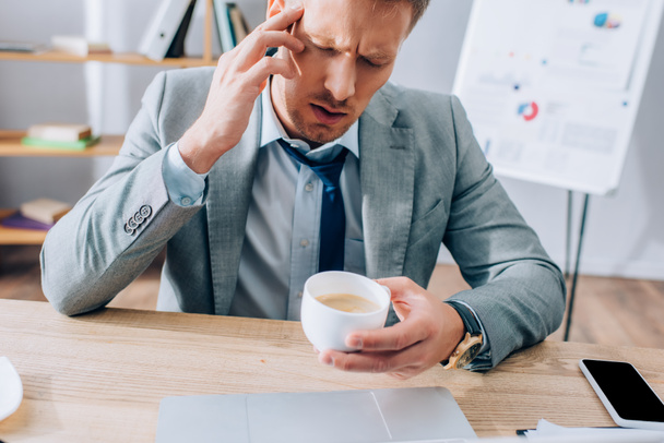 Uomo d'affari che soffre di mal di testa mentre tiene in mano una tazza di caffè vicino a smartphone e laptop in ufficio  - Foto, immagini