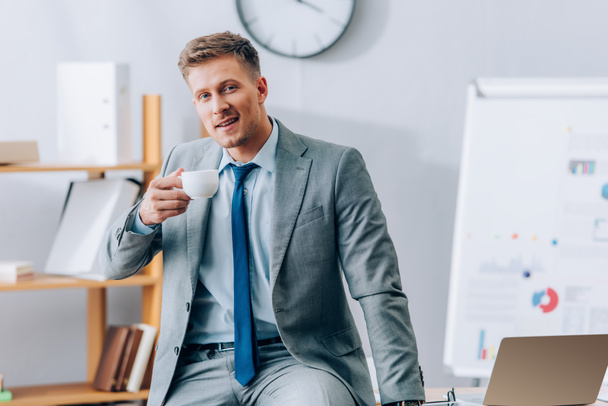 Smiling businessman holding cup of coffee near laptop - Photo, Image