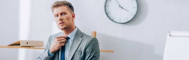 Young businessman adjusting tie and looking at camera in office, banner  - Foto, imagen