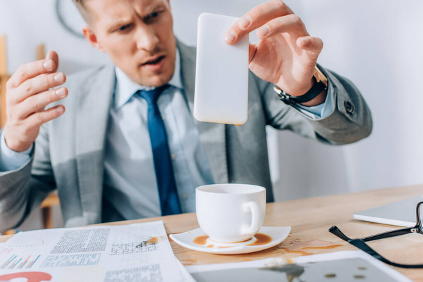 Μπερδεμένος επιχειρηματίας κρατώντας βρώμικο smartphone κοντά ρίχνει έξω καφέ και χαρτιά στο τραπέζι  - Φωτογραφία, εικόνα