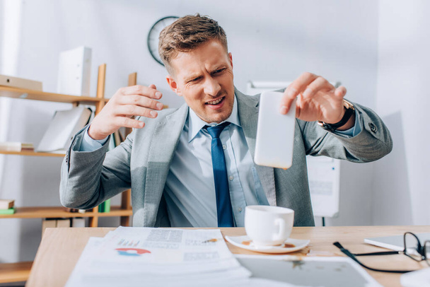 Confused businessman holding dirty smartphone near pouring out coffee on papers and table  - Foto, Imagem
