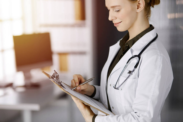 Cheerful smiling female doctor using clipboard in sunny clinic. Portrait of friendly physician woman at work. Perfect medical service in hospital. Medicine concept - Foto, Imagen