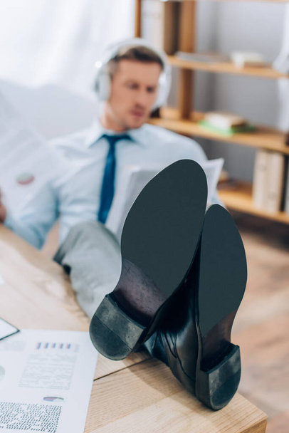 Empresario poniendo piernas en la mesa mientras trabaja con papeles sobre fondo borroso  - Foto, imagen