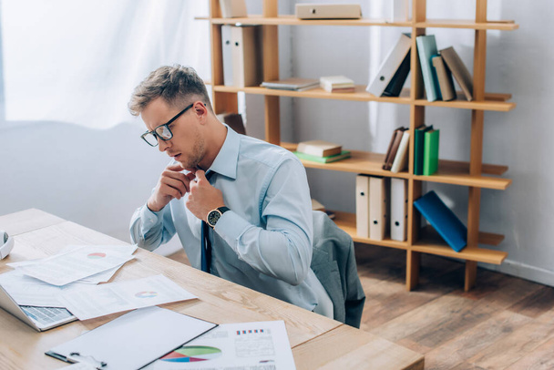 Empresário em óculos tocando gravata enquanto olha para a papelada perto do laptop em primeiro plano borrado  - Foto, Imagem
