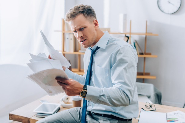 Embarrassed businessman looking at documents near working table on blurred background  - Photo, Image