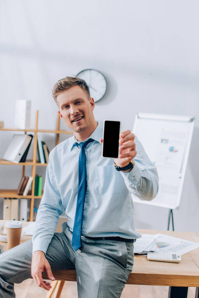 Усміхнений бізнесмен, що показує смартфон з порожнім екраном на розмитому передньому плані в офісі
  - Фото, зображення