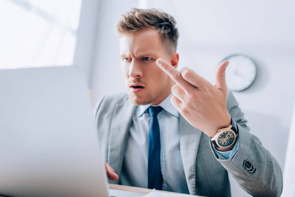 Homem de negócios irritado mostrando o dedo médio ao usar o laptop em primeiro plano desfocado no escritório  - Foto, Imagem