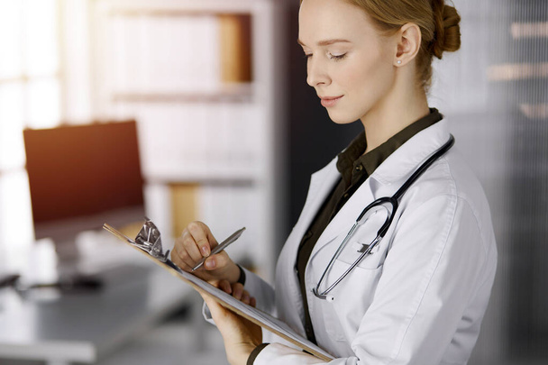 Cheerful smiling female doctor using clipboard in sunny clinic. Portrait of friendly physician woman at work. Perfect medical service in hospital. Medicine concept - Foto, imagen