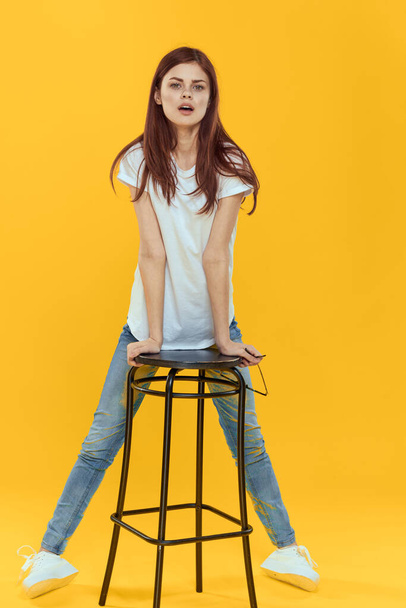 Woman sitting on a chair white t-shirt jeans fashionable clothes glamor studio yellow background - Fotoğraf, Görsel
