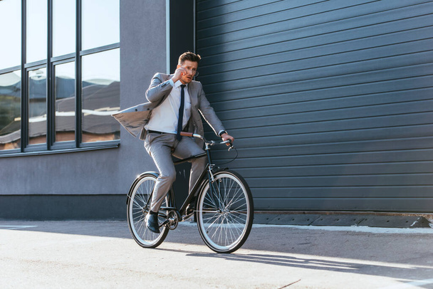 Uomo d'affari in giacca e cravatta che tiene gli occhiali mentre pedala vicino all'edificio  - Foto, immagini