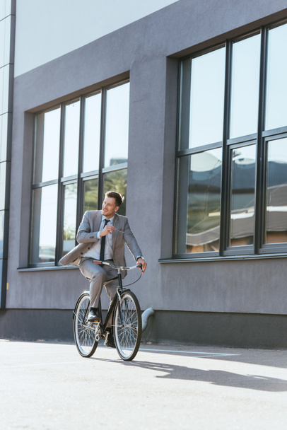 Smiling businessman holding eyeglasses while riding bike outdoors  - Фото, изображение