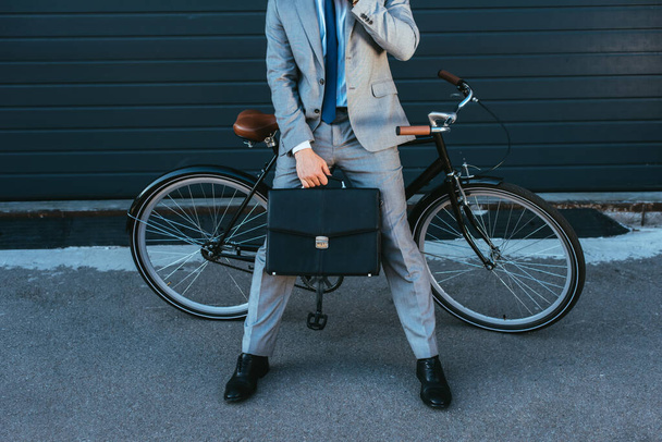Cropped view of businessman holding briefcase while standing near bike and building outdoors  - Photo, image