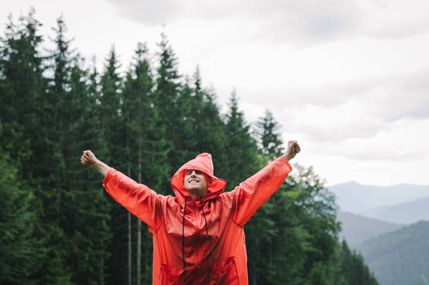 Щасливий і веселий молодий мандрівник у червоному плащі піднімає руки, відчуваючи велике полегшення після сходження на гору. Мальовничий ліс на фоні
. - Фото, зображення