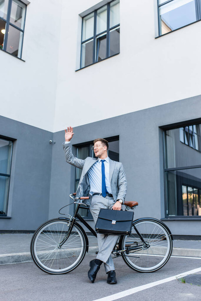 Cheerful businessman waving hand while holding briefcase near bicycle outdoors  - Photo, Image