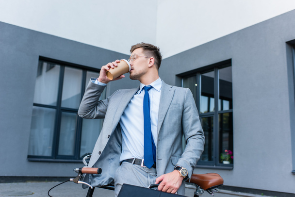 Businessman drinking coffee to go while holding briefcase near bike outdoors  - Photo, Image