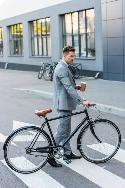 Vista lateral del hombre de negocios sosteniendo café para llevar cerca de la bicicleta en el cruce al aire libre   - Foto, Imagen